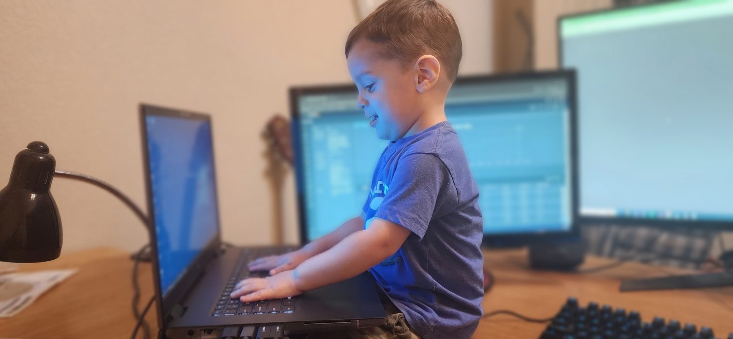 A young child at a laptop at a home desk, focused as if working, with softly blurred monitors behind.