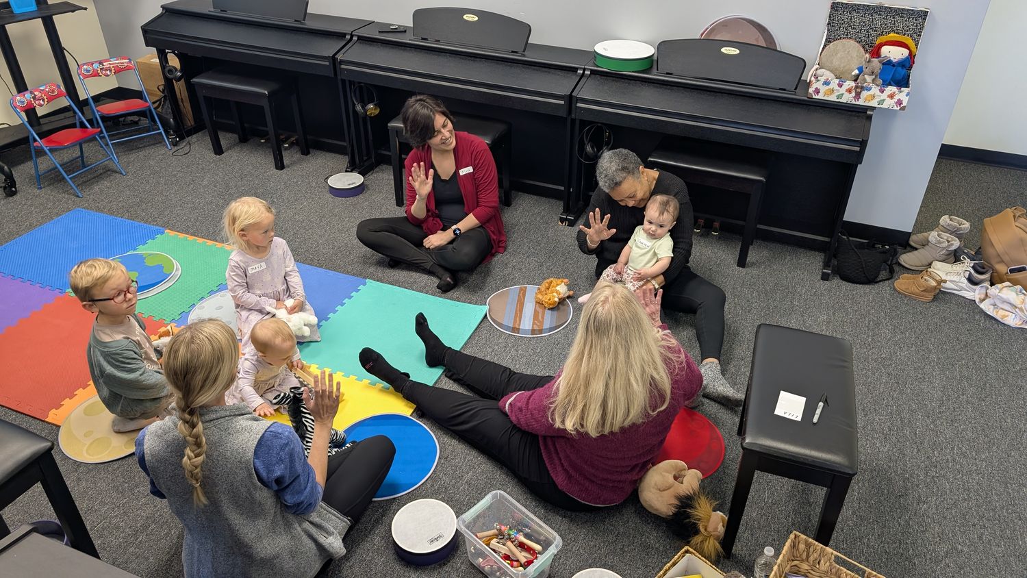 An early-childhood music class sitting in a circle on the floor, teachers and toddlers playing hand instruments together.